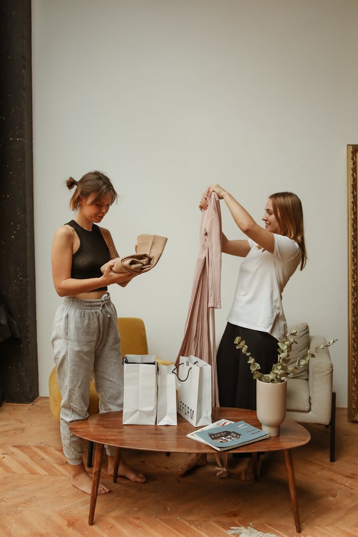 Two young women at home enjoying their new shopping purchases, displaying clothes and gift bags.